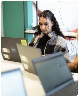 Funcionária Bernhoeft digitando em seu notebook. A foto foi tirada na lateral frontal da mulher. Ela veste blusa preta e casaco quadriculado branco, preto e cinza.