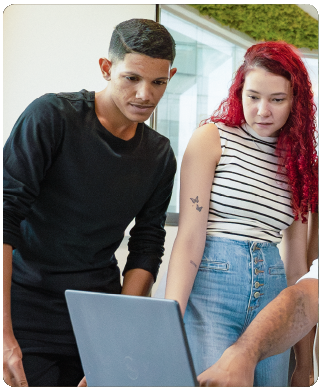 Dois funcionários Bernhoeft em pé olhando para um notebook abaixo deles. A mulher, na direita, é uma mulher branca e ruiva, veste calça jeans e blusa sem mangas com listras pretas. O homem, à sua direita, é um homem negro, veste calça jeans preta e blusa de mangas na cor preta.