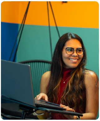 Funcionária Bernhoeft sorrindo para o seu lado. À sua frente há um notebook. Ela é uma mulher negra, com cabelos grandes e ondulados na cor castanho. Veste uma camisa sem mangas na cor vermelha.