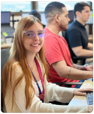 Três funcionários Bernhoeft. Click tirado na paralela. Em primeiro plano, mulher loira sorrindo para câmera. Em segundo plano e desfocado, dois homens.