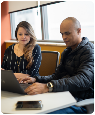 dois funcionários Bernhoeft (um homem e uma mulher) sentados e olhando para o mesmo notebook apoiado à mesa. click com vista de frente para eles.